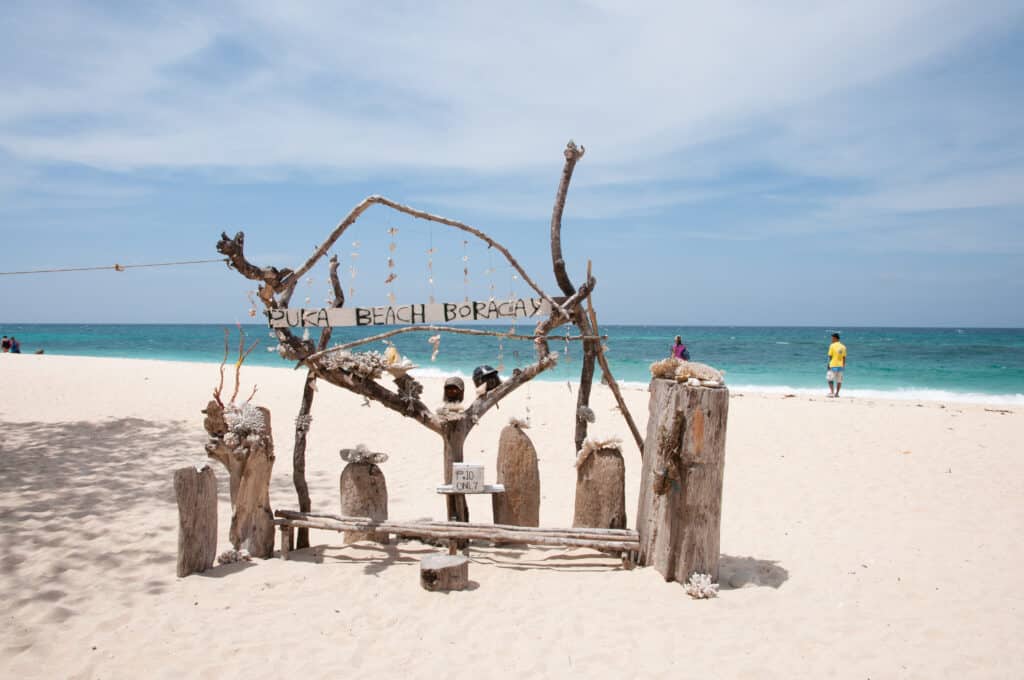 Uma placa rústica de madeira com os dizeres "Puka Beach Boracay" está na areia branca com um fundo de mar e céu azuis. Representa o post de melhores praias nas Filipinas.