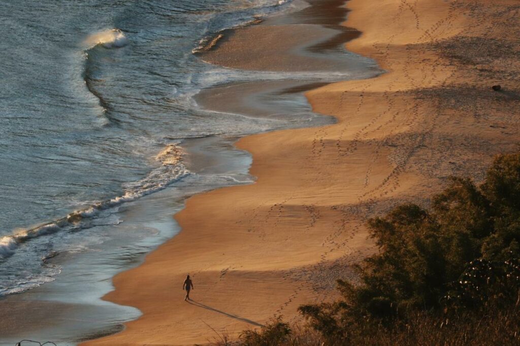 Imagem da praia de Santigo no final do dia com faixa de areia do lado direito com uma pessoa caminhando e do lado esquerdo o mar.