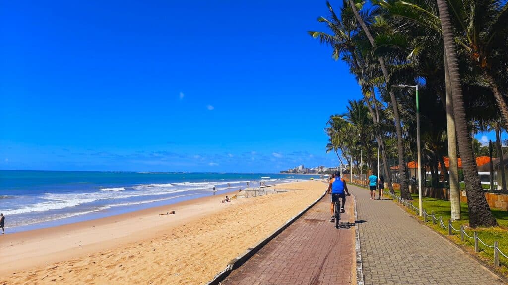 Orla da praia de Jatiúca, com um homem andando de bicicleta, algumas pessoas caminhando na orla, e à esquerda a praia com ondas e faixa de areia branca.
