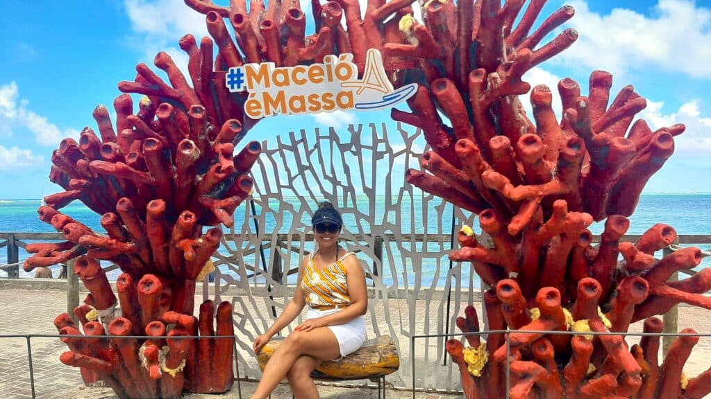 Mulher sentada em um banco, sorrindo e usando boné e óculos de sol, em uma moldura girante vermelha escrita ao topo "Maceió é massa.