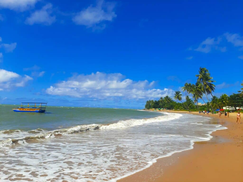 Praia de Sonho Verde com poucas ondas, um barquinho navegando no mar, algumas pessoas andando na areia e coqueiros beirando a orla