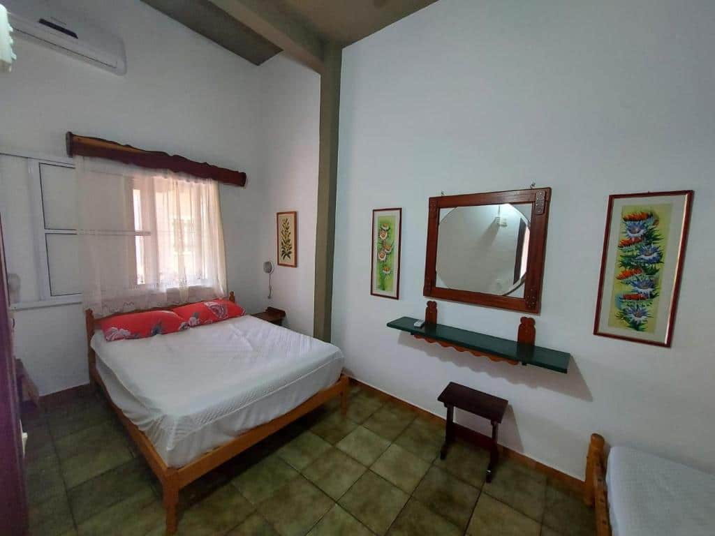 Quarto do Apartamentos Ubatuba com cama de casaldo lado direito da imagem no centro embaixo de uma janela com cortinas, do lado esquerdo da cama a frente uma cômoda com um espelho e um banco. Representa airbnb na praia da Lagoinha.