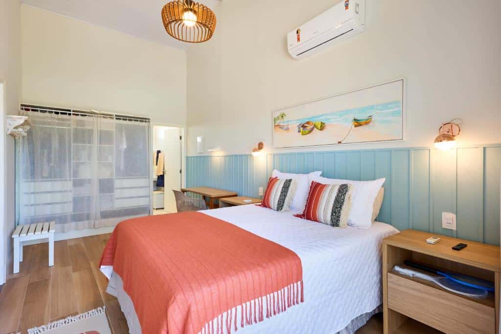 Quarto da Casa de alto padrão com ampla vista para o mar com cama de casal do lado direito da imagem com uma cômoda em cada lado da cama, do lado direito da cama ao lado da cômoda uma mesa com cadeira.