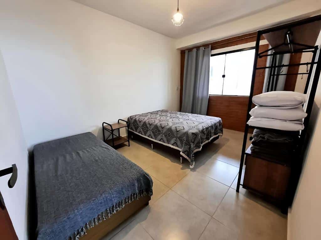Quarto da Casa Amantes da Serra Ibitipoca com duas camas de casal, uma encostada na parede esquerda e outra mais ao centro do quarto. No meio das camas tem uma mesinha e na frente das camas tem uma estante para guardar roupas de cama e objetos. No quarto as paredes são lisas e brancas e tem apenas uma parede do lado direito que é de tijolos e tem uma janela de vidro com cortina.