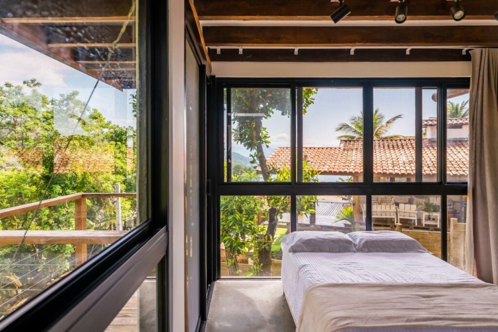 Quarto da Casa Waimea com cama de casal do lado esquerdo da imagem com  uma parede de vidro atrás da cama e do lado direito. Representa airbnb na praia do Félix.