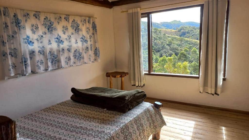 Quarto do Chalé Sabiá com uma cama de frente para uma janela aberta com vista para as montanhas verdes. O chão do quarto é de madeira, tem um banquinho de madeira encostado na parede esquerda e ainda na parede esquerda tem uma janela com cortinas.