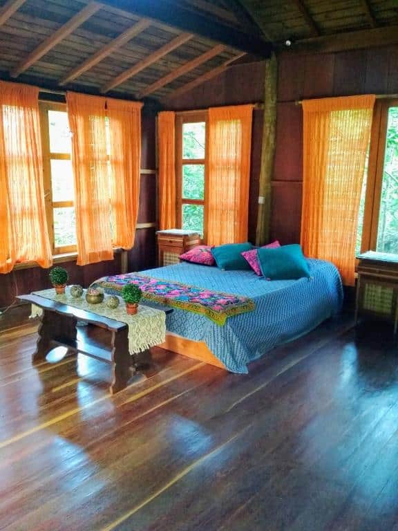 Quarto do Chalé Solar no Matutu com uma cama de casal ao centro e atráa da cama uma parede de madeira com duas janelas grandes cobertas por cortinas. De cada lado da cama tem uma mesa de cabeceira e na frente da cama tem uma mesinha retangular com alguns objetos em cima. O chão do quarto é de madeira e na parede esquerda da cama tem outra janela grande com uma cortina.