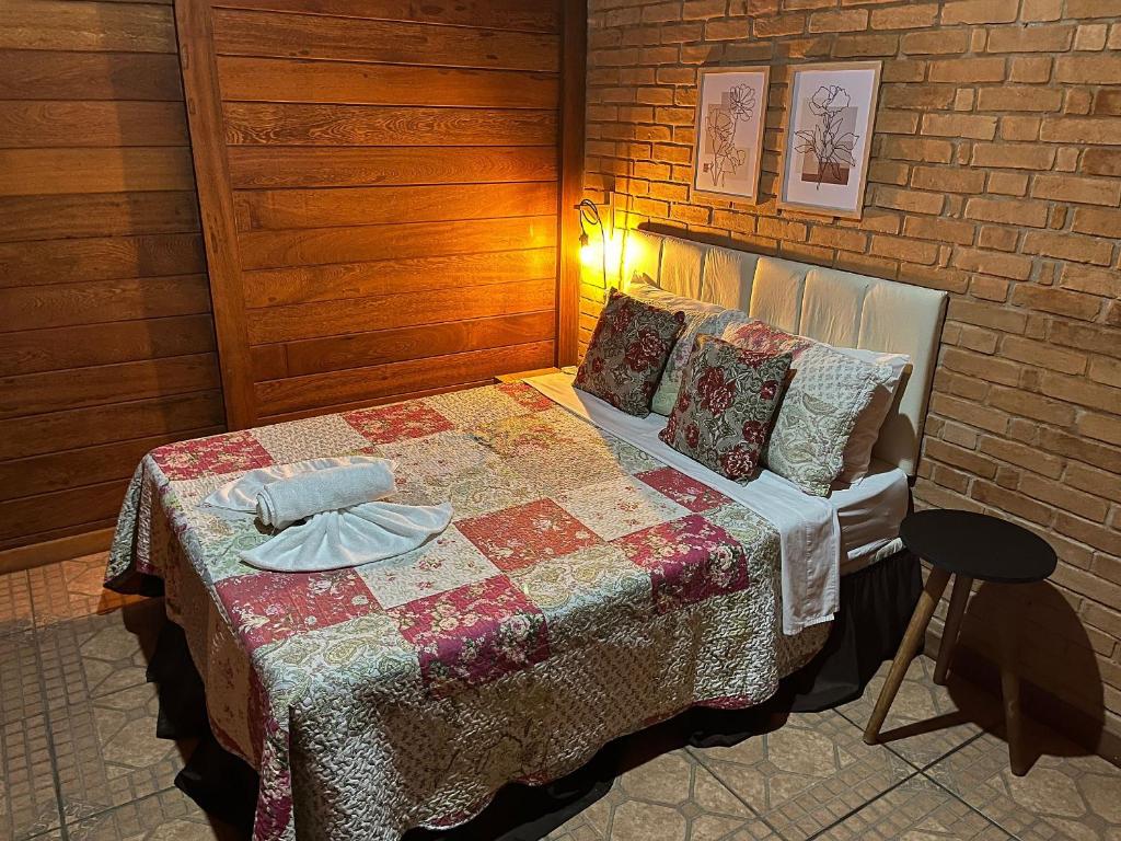 Quarto do Chalés Belo Vale - Rua do Curro com uma cama de casal ao centro, do lado esquerdo tem uma mesinha redonda e do lado direito tem outra mesinha com um abajur. A parede atrás da cama é de tijolos e do lado da cama tem uma parede de madeira.