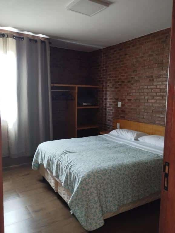 Quarto do Chalés Estrada Real com uma cama de casal ao centro, atrás da cama uma parede de tijolos e ao lado uma estantes para guardar roupas e objetos. O quarto tem uma janela com cortinas e um chão de madeira.