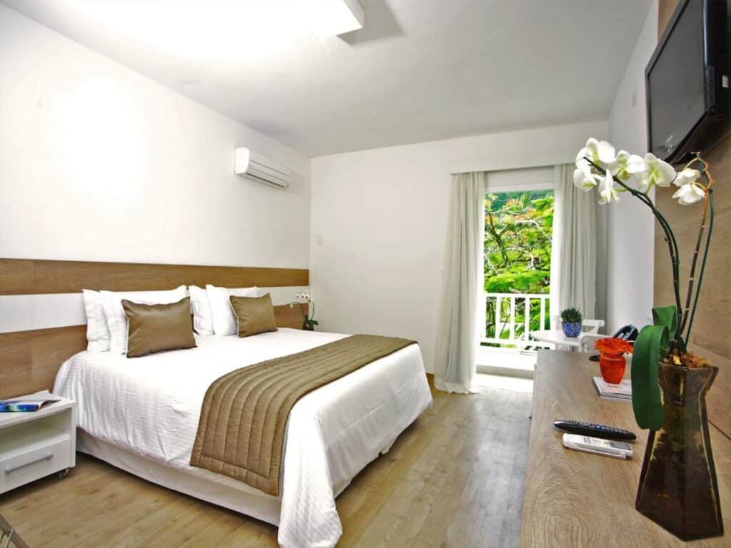 Quarto da Costa Verde Tabatinga Hotel com cama de casal do lado esquerdo da imagem no centro do quarto com uma cômoda em cada lado da cama e em frente a cama uma cômoda de madeira.