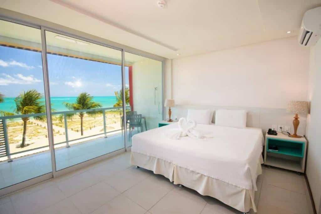 A imagem mostra o interior do quarto do Hotel Praia Dourada com uma grande janela de vidro que oferece uma vista espetacular para uma praia. A cama de casal está centralizada na imagem, com roupa de cama branca e uma toalha dobrada em forma de cisne sobre ela. Ao lado da cama, há um criado-mudo com uma lâmpada de base azul e uma tela. Uma pequena mesa e cadeira estão posicionadas próximas à janela, ideal para apreciar a vista ou para uma refeição leve. O chão é de azulejo claro, e a decoração é minimalista, focando na paisagem deslumbrante vista através da janela, onde a praia de areia branca se encontra com o mar azul-turquesa e é ladeada por palmeiras.