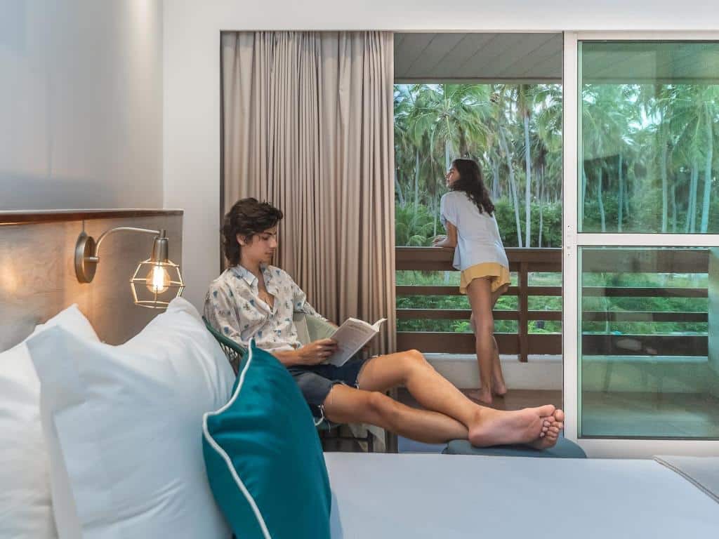 Foto do Quarto do Japaratinga Resort. À esquerda, um jovem está sentado na cama, lendo um livro. Ele está relaxado, vestindo uma camisa estampada e shorts, com os pés descalços apoiados na cama. A cama é grande, com roupa de cama branca e travesseiros confortáveis. Ao lado da cama, há uma luminária de mesa moderna com uma base de metal.
À direita, uma mulher está em pé na varanda que é acessível através de uma grande porta de correr de vidro. Ela está de costas para a câmera, olhando para fora, vestida com uma camiseta branca e shorts amarelos. A varanda tem uma vista direta para um cenário tropical, com palmeiras e outra vegetação exuberante. O quarto é iluminado e arejado, com uma sensação de serenidade e conexão com a natureza.