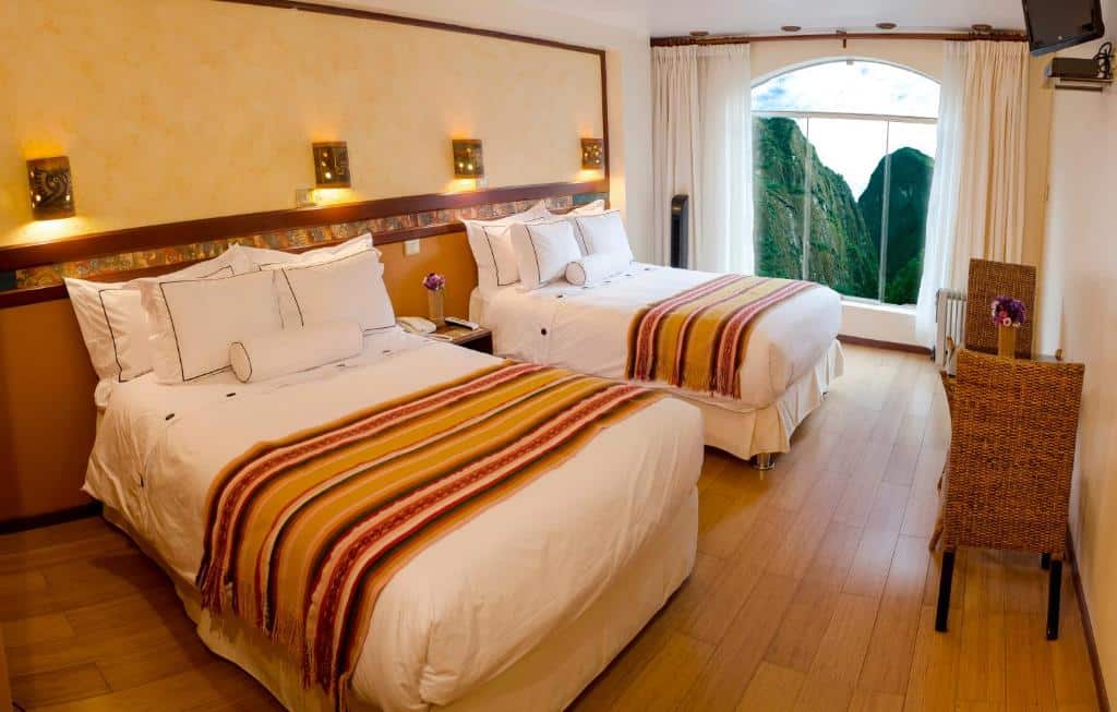 suíte do hotel taypikala machupicchu com duas camas de casal lado a lado, uma janela grande com vista para as montanhas e uma pequena mesa de canto com duas cadeiras de vime