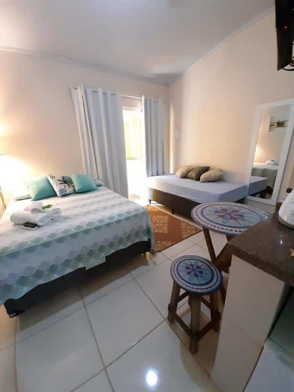 Airbnb Flats Jordão. No canto esquerdo há uma cama de casal, uma janela no meio e no lado direito há uma cama de solteiro. Ainda no canto direito há uma mesa pequena com dois banquinhos. Imagem para ilustrar o post airbnb em Ilha Grande.