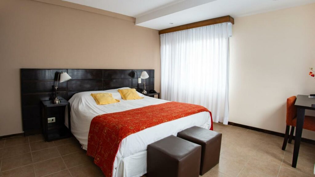 Quarto do Hotel Poincenot  com cama de casal do lado esquerdo da imagem no centro em cada lado da cama uma cômoda com luminária e no pé da cama dois puffs. Representa hotéis em El Chalten.