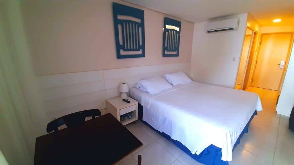 Interior de um dos quartos do Hotel Ponta Verde Francês. O local tem uma mesinha quadrada com duas mesas, uma cama de casal com uma escrivaninha ao lado e ar-condicionado