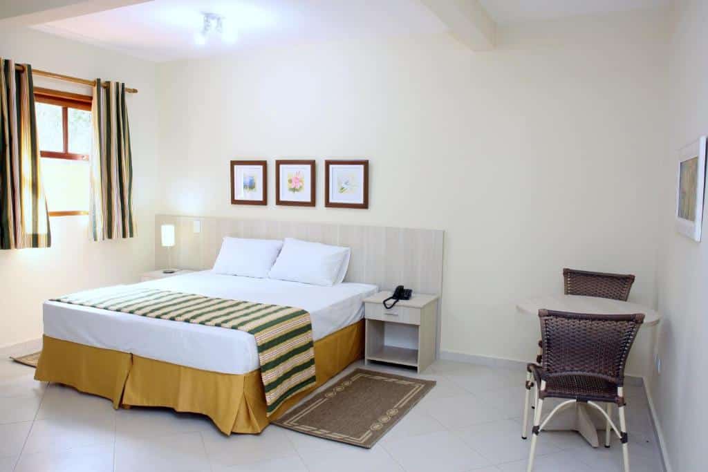 QUarto do Hotel Refúgio Vista Serrana com uma cama de casal, onde há uma cabeceira e duas mesinhas de cabeceira, do lado esquerdo da cama há uma janela com cortinas, e do lado direito há uma mesa redonda com duas cadeiras