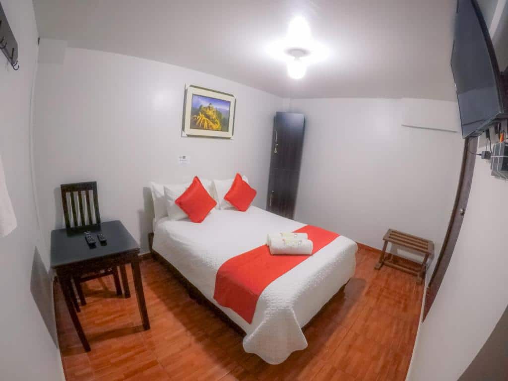 quarto do machu picchu land com uma cama de casal ao centro, decorada com lençóis laranja e branco, um quadro acima da cabeceira, e uma mesa de madeira escura e simples no lado esquerdo da cama,