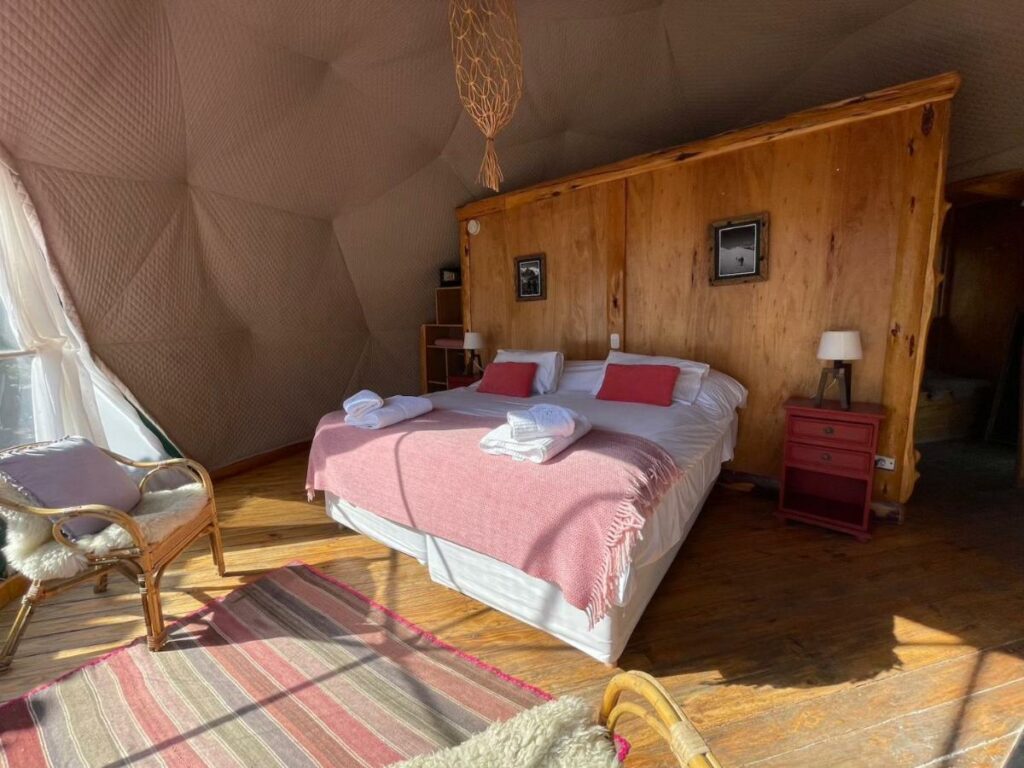 Quarto do Patagonia Eco Domes com cama de casal do lado direito da imagem com uma cômoda em cada lado com luminária e em frente a cama do lado direito da cama uma poltrona.
