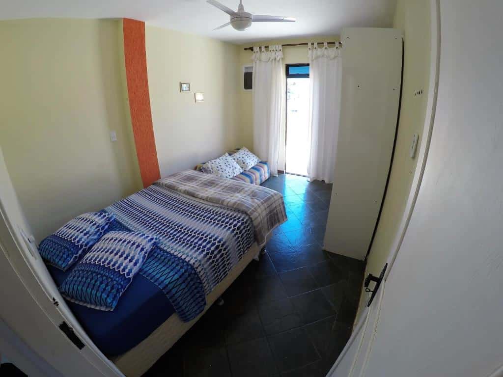 Quarto do airbnb Praia Linda Hospedagem. No lado esquerdo há uma cama de casal, no fundo do quarto há um pequeno sofá no lado esquerdo e um armário no lado direito e ainda no fundo há uma porta que dá acesso ao terraço do apartamento. Imagem utilizada para ilustrar o post airbnb em Arraial do Cabo.