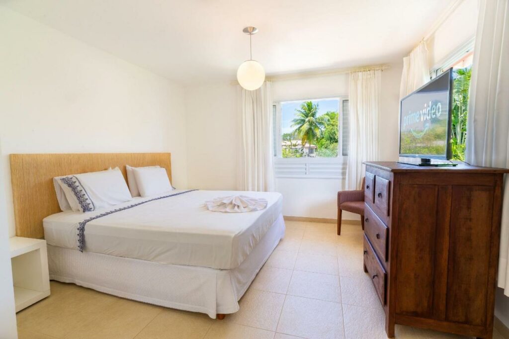 Quarto do Quintas de Sauípe – Casa D13 com cama de casal do lado esquerdo da imagem, em frente a cama uma cômoda com TV. Representa airbnb na Costa do Sauípe