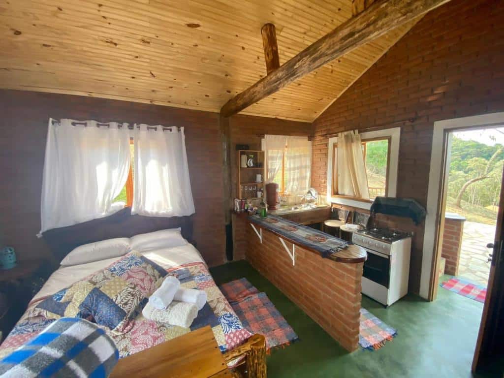 Parte interna do chalé RECANTO DO LEÃO que mostra na parte direita da imagem uma cozinha compacta com fogão a gás, duas banquetas, uma pia, uma prateleira com utensílios e uma bancada de tijolos. Na parte da cozinha tem duas janelas, uma em frente a pia e outra atrás das banquetas. Já do lado esquerdo tem uma cama de casal com uma janela atrás e um tapete do lado direito da cama. As paredes são de tijolos, o teto de madeira e do lado do fogão tem uma porta de entrada.