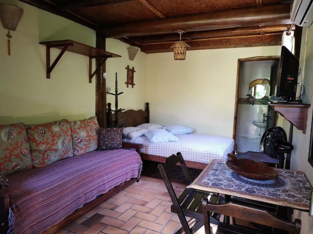 Sala de estar do Recanto Tangará – Chalé – Praia do Felix com mesa com duas cadeiras do lado direito, do lado esquerdo um sofá cama e ao do lado esquerdo do sofá uma cama de casal. Representa airbnb na praia do Félix.