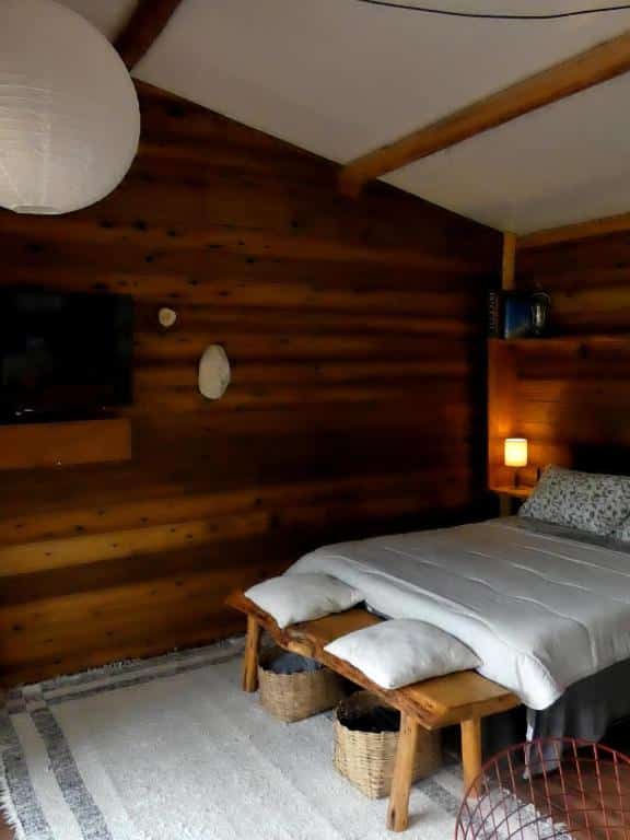 Quarto do Reserva Jacarandá Cabanas com uma cama de casal e no pé da cama tem um banco de madeira com duas almofadas e com dois cestos e um tapete embaixo. As paredes do quarto são de madeiras e tem uma tv na parede.