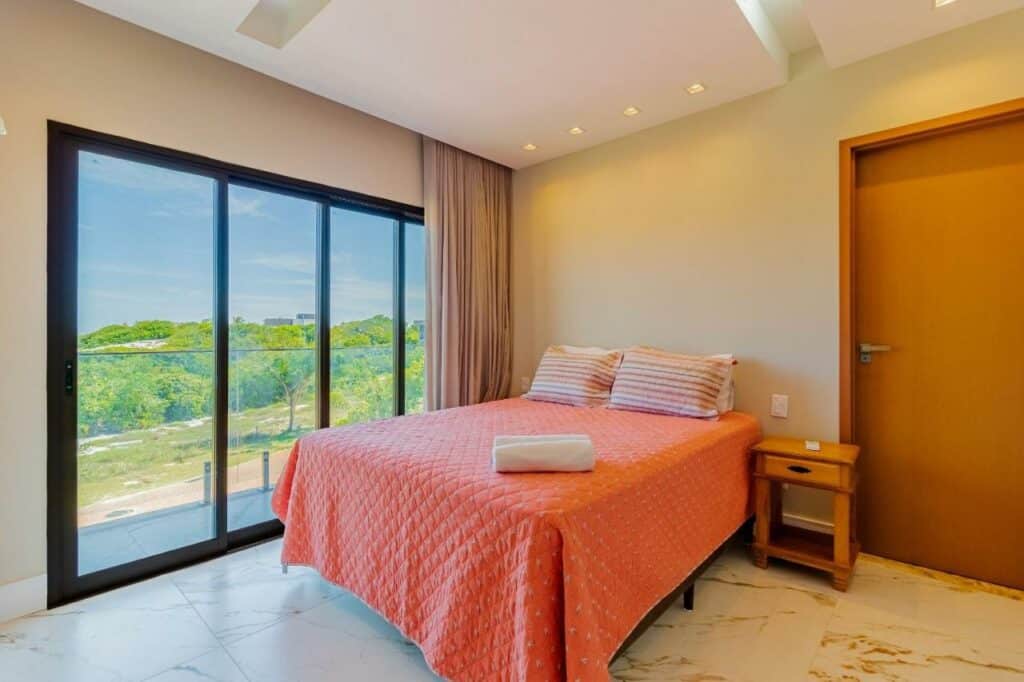Quarto do SA07 Maravilhosa Casa 5 Suítes – Reserva de Sauípe com cama de casal do lado esquerdo da imagem e do lado esquerdo da cama uma cômoda e do lado direito da cama porta de vidro que dá acesso a varanda. Representa airbnb na Costa do Sauípe