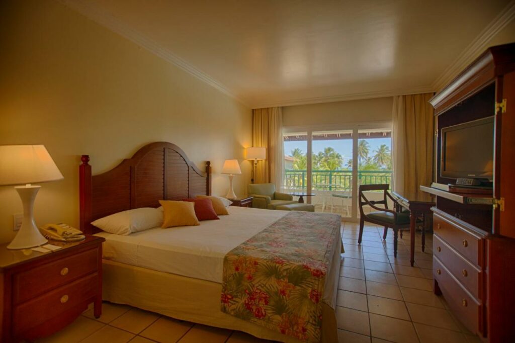 Quarto do Sauipe Resorts Ala Mar - All Inclusive com cama de casal do lado esquerdo da imagem com uma comoda em cada lado da cama com luminária, em frente a cama uma cômoda com TV e do lado esquerdo da cama um sofá ao fundo e em frente ao sofá uma mesa de madeira com cadeira. Representa airbnb na Costa do Sauípe