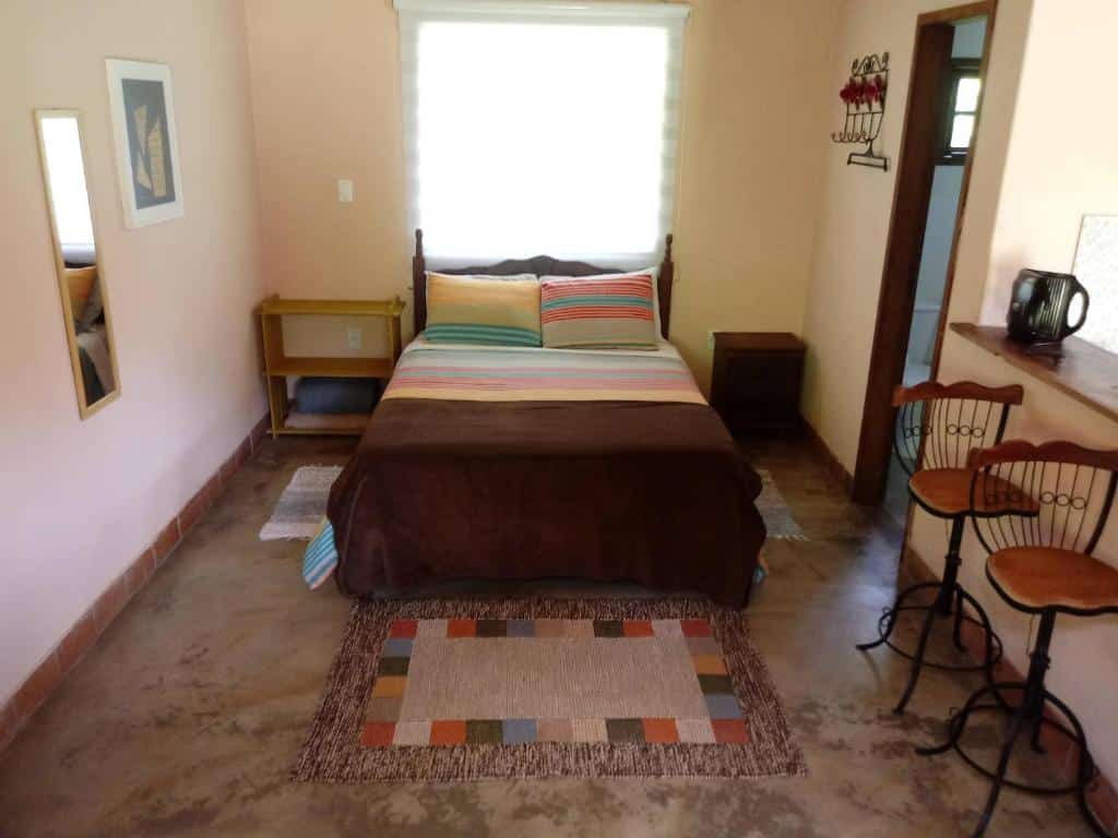 Quarto de 30 m² do Sítio das Hortênsias Chalés com uma cama de casal ao centro, uma janela atrás da cama, uma mesinha de cada lado da cama e um tapete de cada lado da cama. Na parede esquerda do quarto tem um quadro e um espelho, já na parede direita tem uma porta que dá acesso ao banheiro e uma bancada de madeira com duas banquetas que separa a cozinha do quarto.