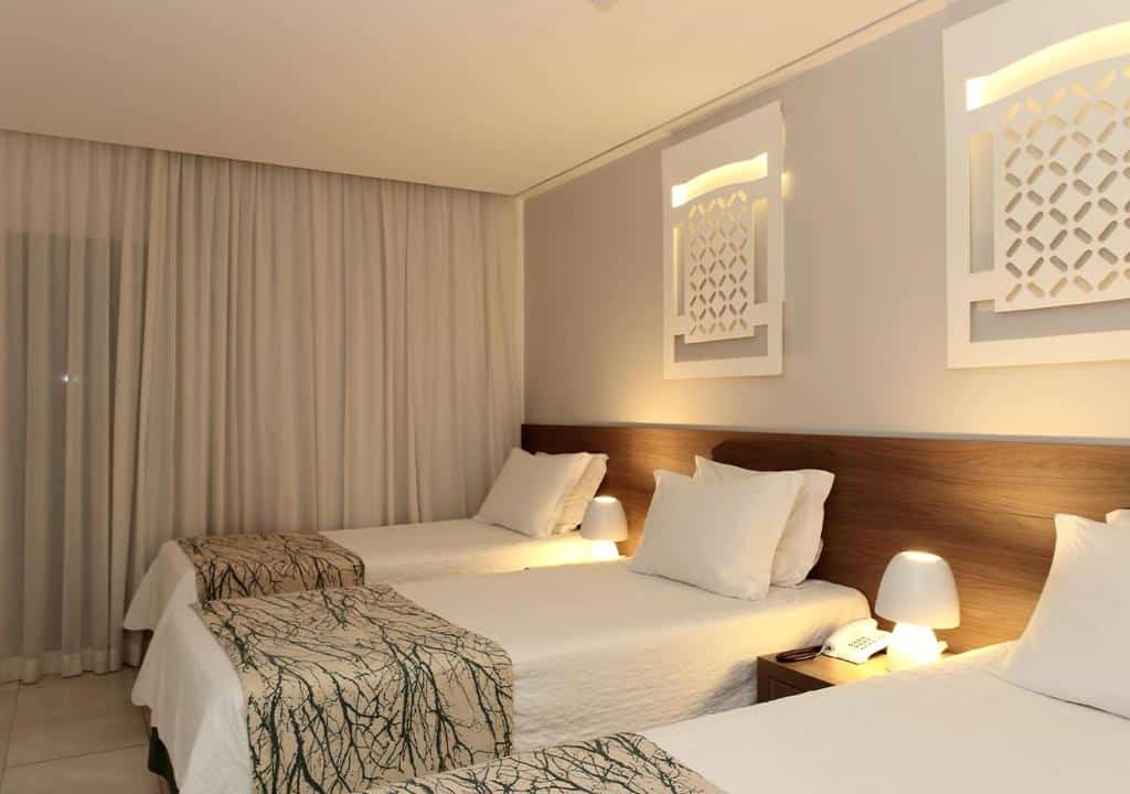 Quarto Standard Triplo do Hotel Ponta Verde Francês. Há três camas com escrivaninha ao lado com luminária em cima acesa e uma cortina blackout fechada