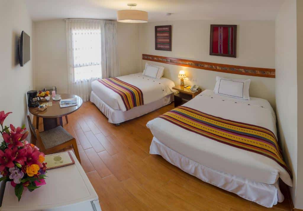 quarto duplo do Taypikala Hotel Cusco com duas camas de solteiro, uma mesa redonda de madeira e duas cadeiras. Em cima da mesa há uma cafeteira elétrica e algumas frutas.