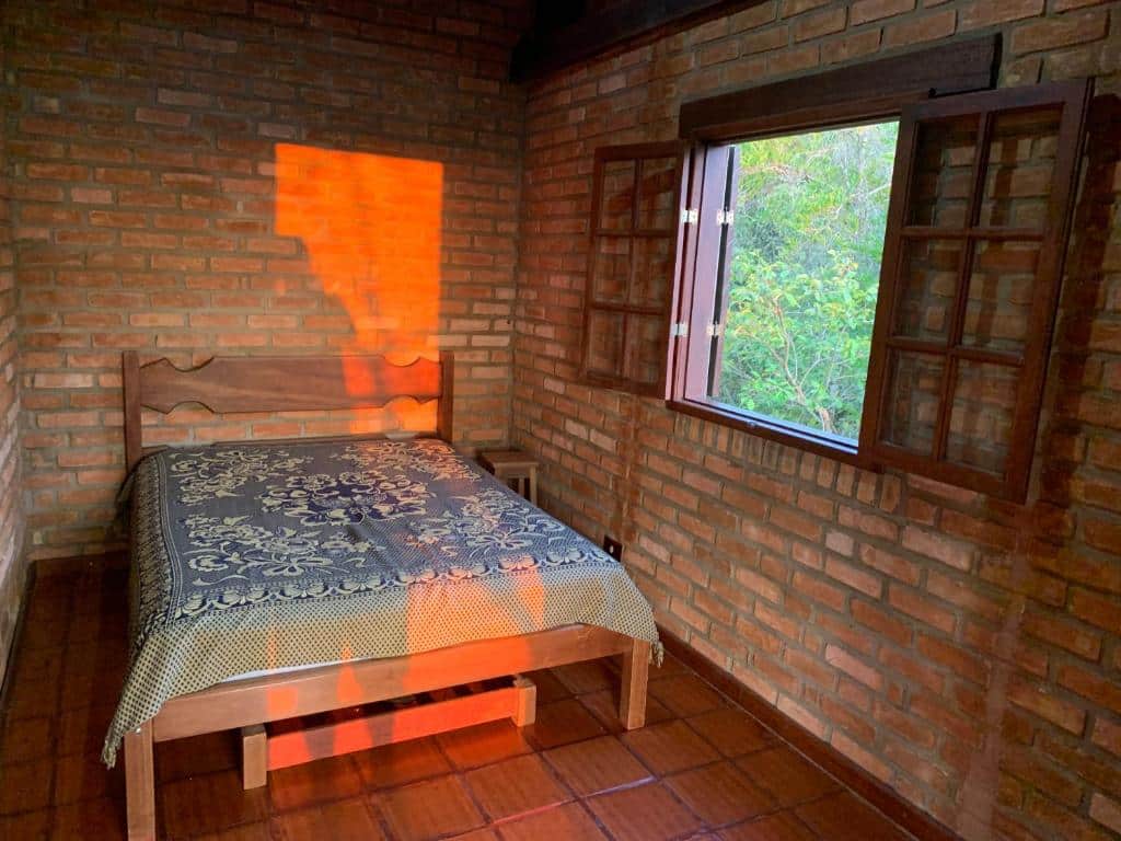 Quarto do Varandas da Serra / Reserva Alto da Colina com uma cama de casal ao centro e uma mesinha do lado direito. As paredes do quarto são de tijolos, o chão é de azulejos e na parede direita tem uma janela aberta.