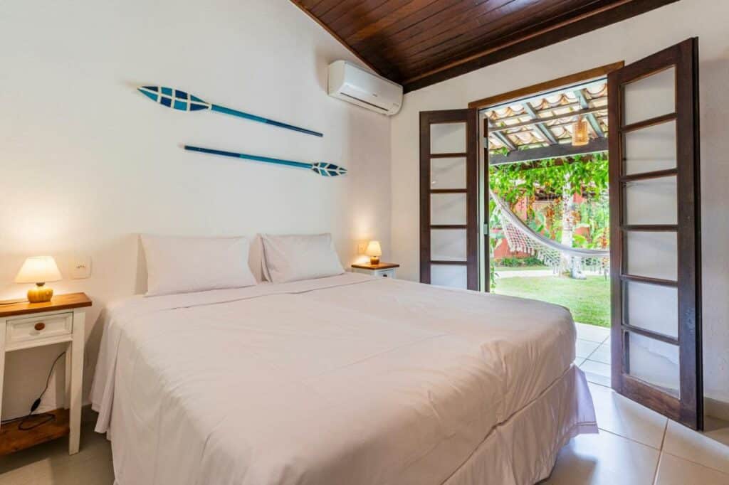 Quarto da Vila Damai Maresias do lado esquerdo da imagem no centro do quarto com uma cômoda em cada lado com luminária e do lado esquerdo da cama uma porta que dá acesso a varanda térrea. Representa airbnb na praia da Juréia.