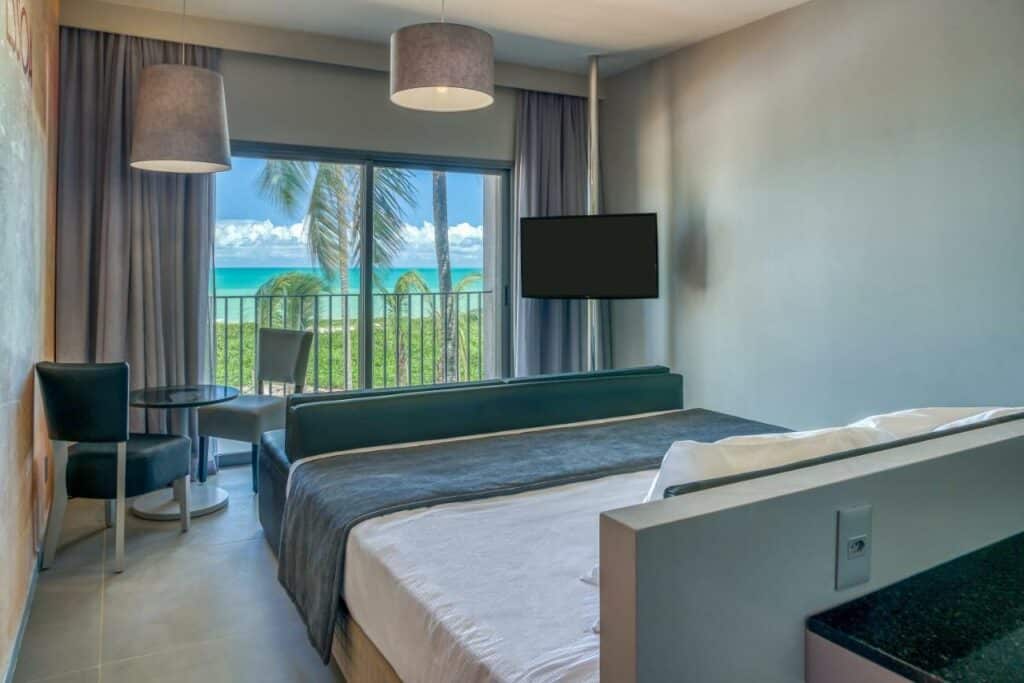 Quarto do Vila Galé Resort Alagoas. Na imagem há uma cama de casal com coberta dobrada em cima na beira da cama, uma TV, uma mesa redonda com duas cadeiras e uma porta de vidro com vista da varanda e do mar. Há um gramado entre a varanda e a praia, e vários coqueiros