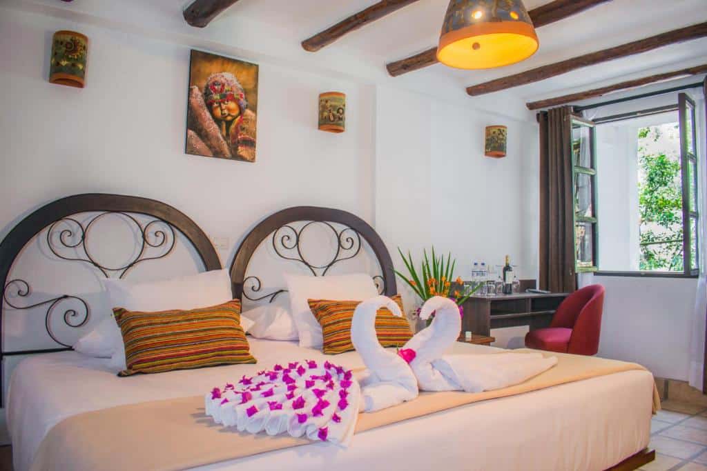 cama de casal no vilas insight hotel machupicchu com duas toalhas ornamentadas como cisnes e decoradas com pétalas de rosas cor de rosa, além de uma pequena mesa de madeira no canto direito e uma janela ao lado que oferece bastante luz natural.