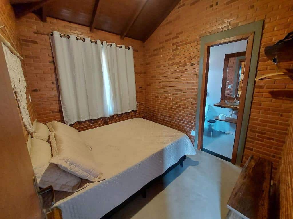 Quarto do Villa da Serra chalé com uma cama de casal com travesseiros e uma colcha e paredes de tijolos. Na parede do lado esquerdo da cama tem uma janela com cortina, na parede em frente a cama tem uma porta aberta que dá acesso ao banheiro, e na parede direita tem um banco de madeira encostado.