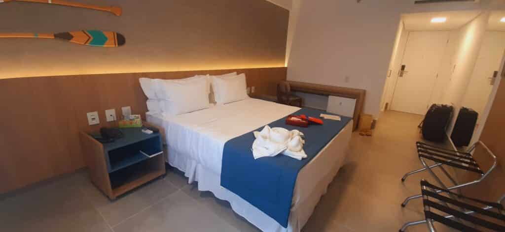 Quarto Vista Piscina do Maragogi Exclusive Hotel, com uma cama de casal com travesseiros e mimos em cima, uma mesinha ao lado direito e, no lado esquerdo, tem um balcão com cadeira e um frigobar embaixo.