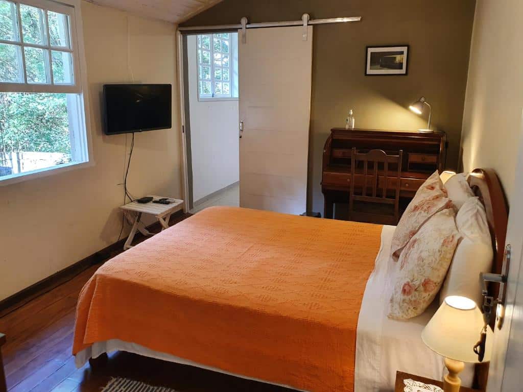 Quarto do airbnb Rancho São Carlos. No fundo da imagem está a porta do quarto no lado esquerdo e uma mesa e cadeira no lado direito. Já no lado esquerdo há uma janela com vista para área verde e uma TV presa a parede com um pequeno banco embaixo. Na parede do lado direito está encostada a cama de casal. Imagem para ilustrar o post airbnb em Itatiaia.