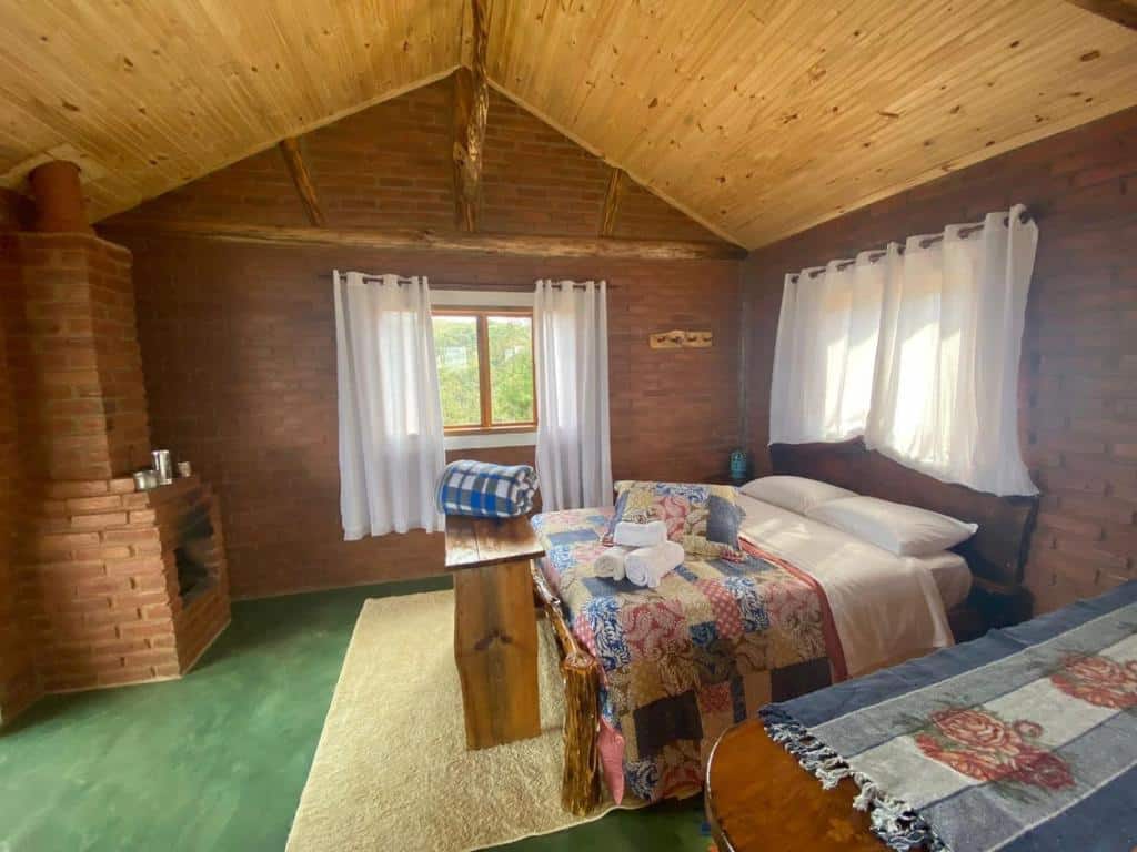 Quarto do RECANTO DO LEÃO que mostra uma cama de casal do lado direito do quarto com uma janela atrás e na frente da cama uma bancada de madeira. Já do lado esquerdo do quarto tem uma lareira de tijolos e ao fundo na parede outra janela. As paredes dos chalés são de tijolos e o teto é de madeira. Imagem ilustrando post Airbnb em Aiuruoca.