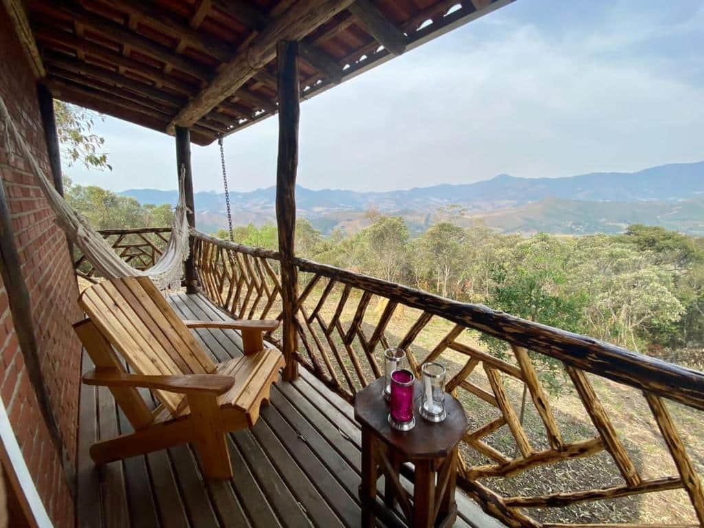Varanda com chão de madeira do chalé RECANTO DO LEÃO. Na varanda tem uma poltrona de madeira, uma rede ao fundo e um banquinho com três copos. A varanda tem telhado de telhas e a proteção é de madeiras. Já a vista da varanda dá para as árvores e montanhas durante o dia, ilustrando post Airbnb em Aiuruoca.