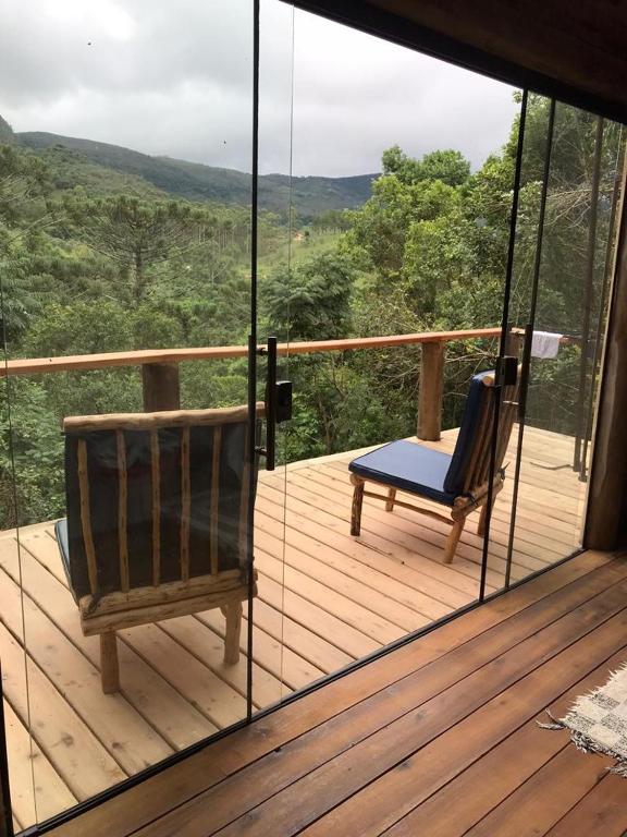 Parte do Reserva Jacarandá Cabanas que mostra a varanda do quarto com uma porta de vidro grande que dá acesso a varanda com chão de madeira e duas poltronas com vista para as árvores e montanhas durante o dia. Imagem ilustrando post Airbnb em Aiuruoca.
