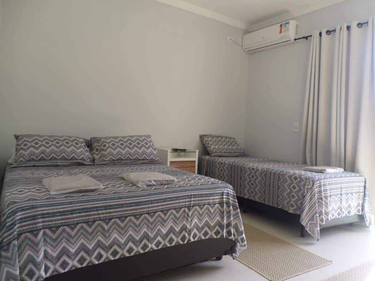 Quarto do Residencial Camburyá, um dos airbnb em Camburizinho. Uma cama de casal e uma de solteiro tem uma mesinha de cabeceira entre si. Na parede ao lado da cama de solteiro há uma janela com cortinas, e logo ao lado fica um aparelho de ar-condicionado.