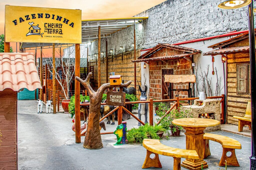 Restaurante Cheiro da Terra, em Maceió. A foto é da área externa, onde tem uma placa escrito "Fazendinha Cheiro da Terra" e uma área com bancos, flores, réplicas grandes das casas do sertão e um parquinho ao fundo