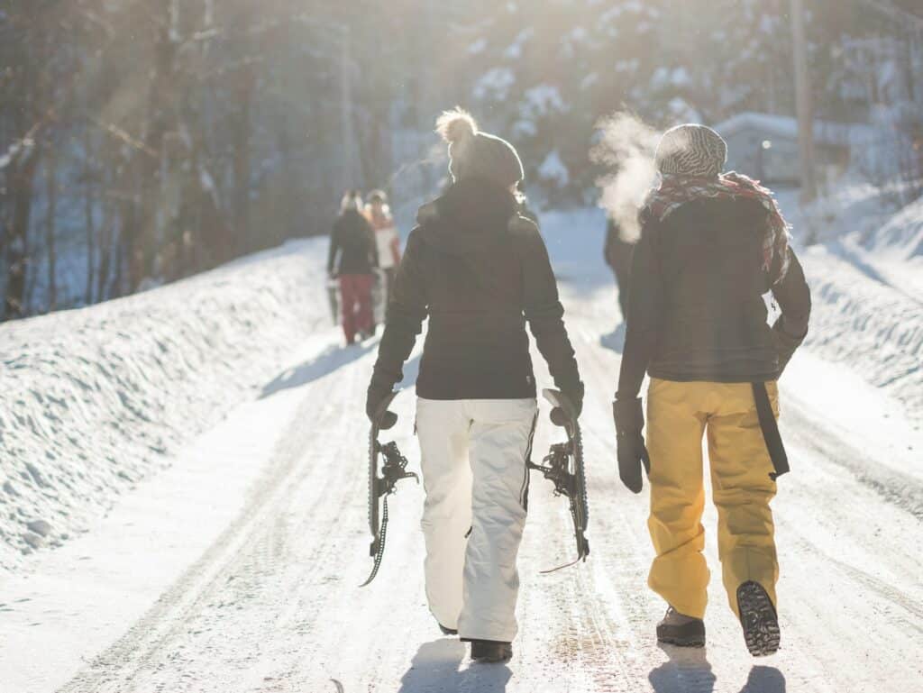vista de pessoas de costas em meio à neve carregando esquis, caminhando e conversando animadas em uma estrada com árvores nos arredores em Saint-Adolphe-d'Howard, no Canadá