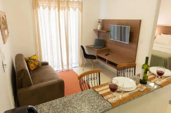 Sala de estar do airbnb Apart Sol Nascente. No lado esquerdo há um sofá, no fundo da sala há uma cortina grande e no lado direito há um rack com uma TV e um notebook, na frente do rack há uma cadeira. Já na frente da imagem há uma bancada que divide a sala da cozinha e em cima dela há dois pratos com talheres, taças de vinho e uma garrafa de vinho. No canto direito da imagem é possível ver uma parte do quarto por uma porta que está aberta.