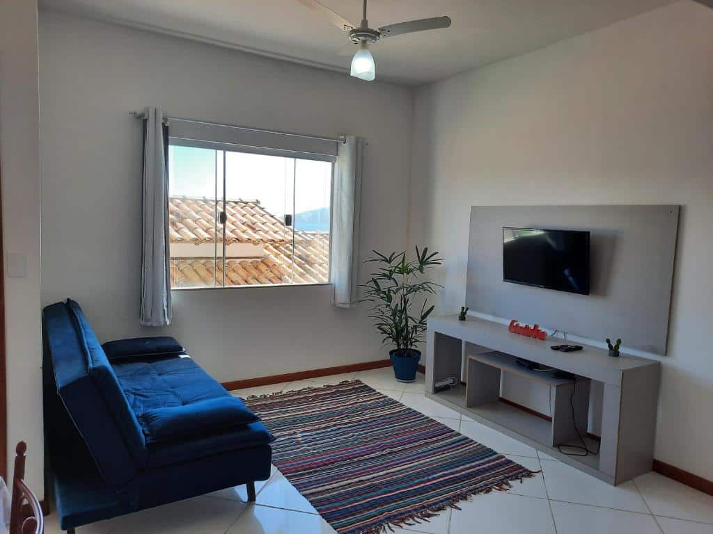 Sala de estar do airbnb Bela Vista Flats. No lado esquerdo da sala está o sofá, no meio da sala há um tapete no chão e no lado direito há um rack com uma TV presa. No lado esquerdo do rack há uma planta e uma janela de vidro.