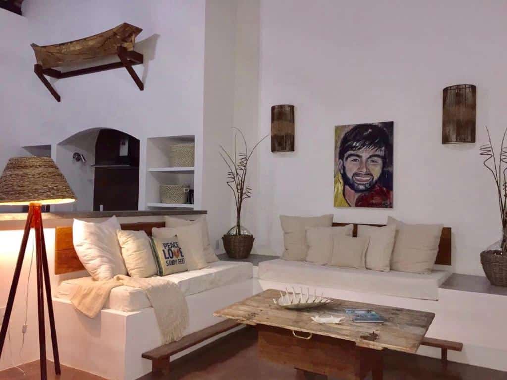 Sala de estar do airbnb Casarao da Praia. O sofá possui formato em L, no meio do cômodo a uma mesa de centro e o cômodo é dividido por uma bancada no fundo que dá acesso a cozinha.
