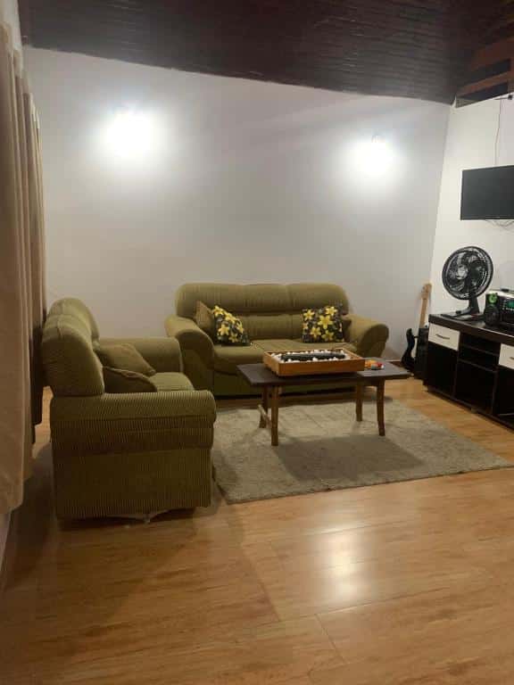 Sala de estar do airbnb Chalé no centro de Penedo. No lado direito há um rack com um ventilador em cima e uma TV presa a parede, no meio da sala há uma mesa de centro e um sofá atrás e no lado esquerdo há outro sofá e uma cortina atrás do sofá.